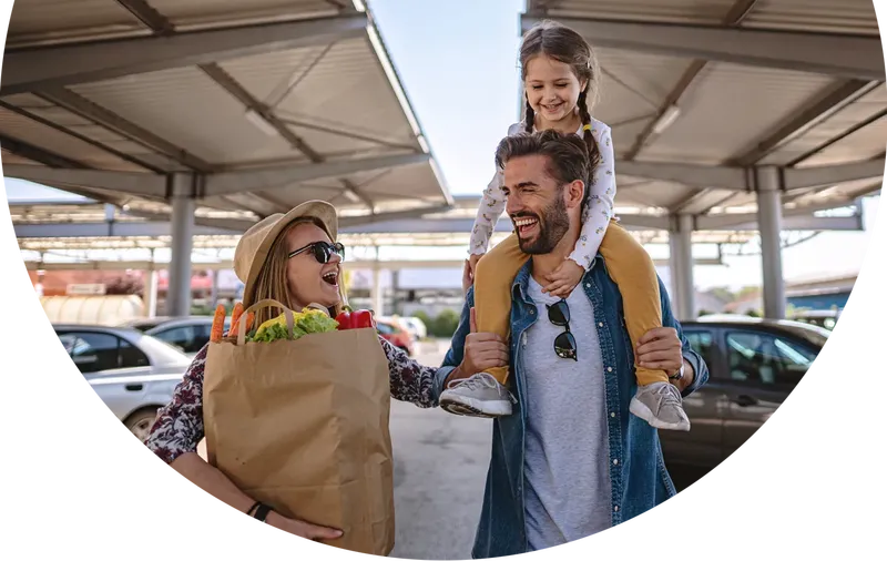Eine Familie geht nach dem Einkauf über den Parkplatz. Der Vater hat die Tochter auf den Schultern sitzen und die Mutter trägt die Einkaufstüte. Alle lachen.