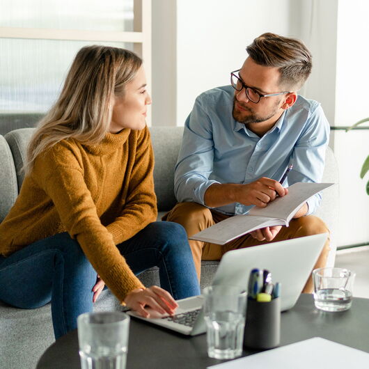 Eine Frau und ein Mann sitzen vor einem Laptop und schauen sich Unterlagen an