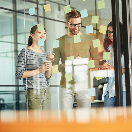 Drei Kollegen stehen vor einer Glaswand, an die sie Klebenzettel kleben