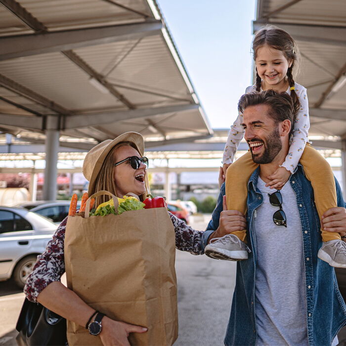 Eine Familie geht nach dem Einkauf über den Parkplatz. Der Vater hat die Tochter auf den Schultern sitzen und die Mutter trägt die Einkaufstüte. Alle lachen.